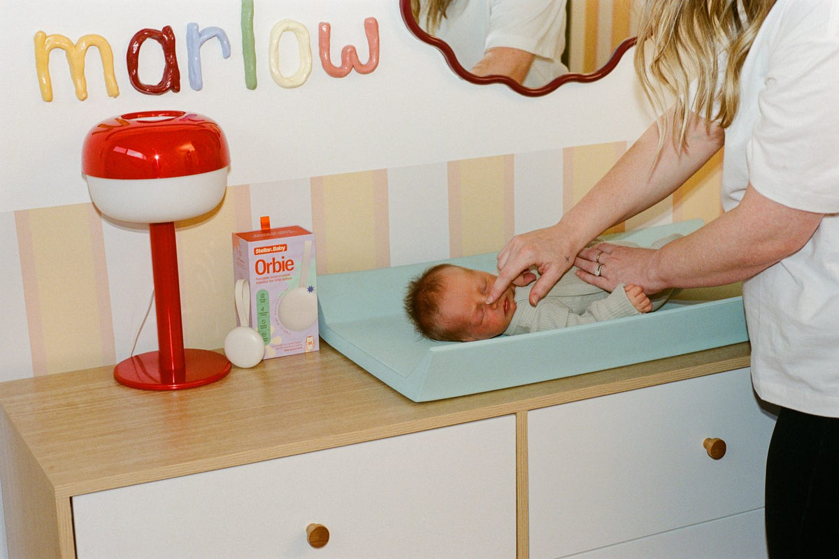 A parent changing a baby beneath a name sign, with Orbie and its box on the drawers