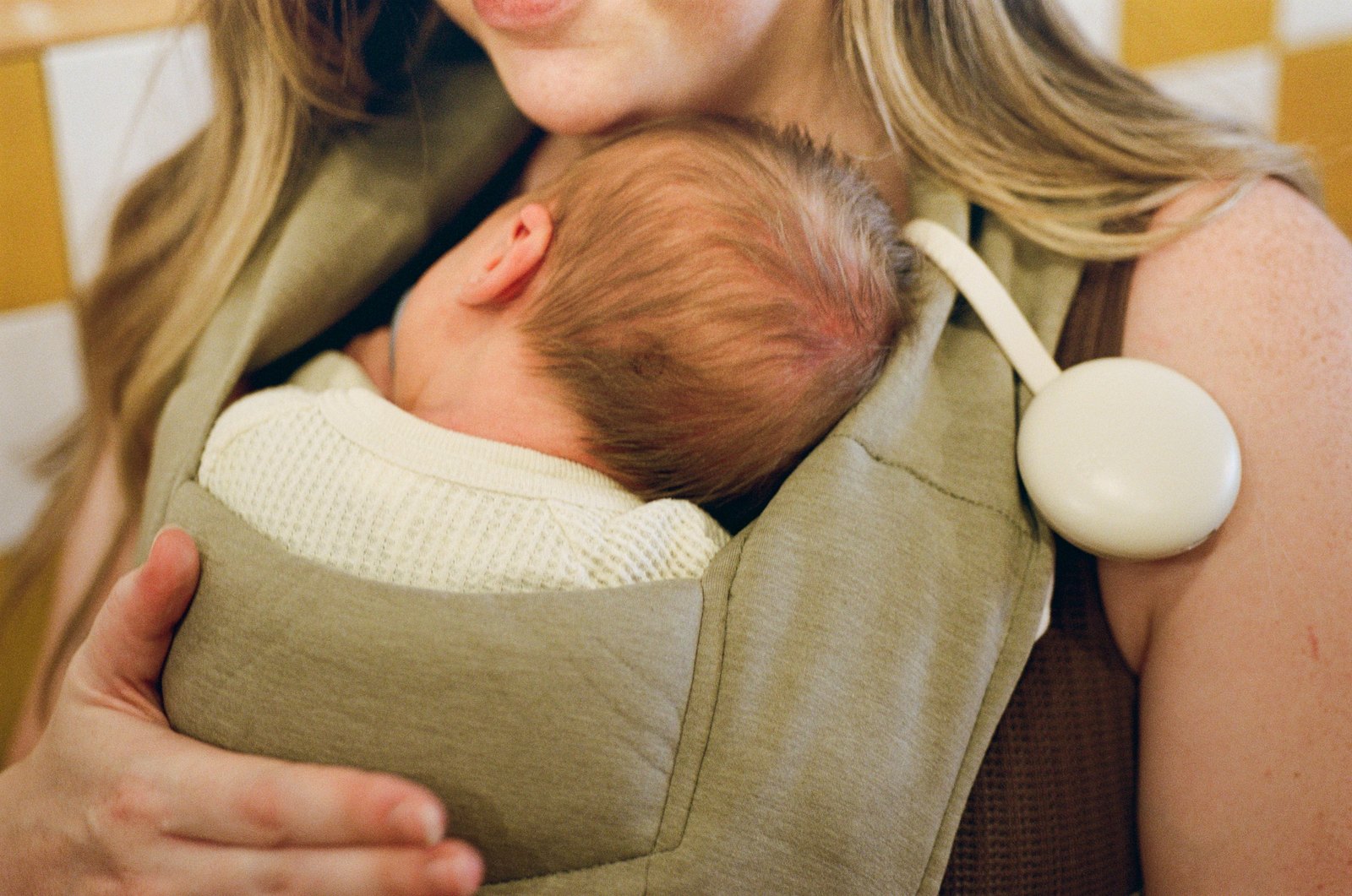 A newborn in a carrier against a parent’s chest, with Orbie clipped alongside